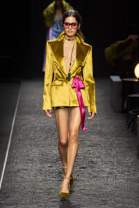Model walks the runway in a metallic gold satin blazer mini dress with a bright pink ribbon belt and pink sunglasses, heels included.