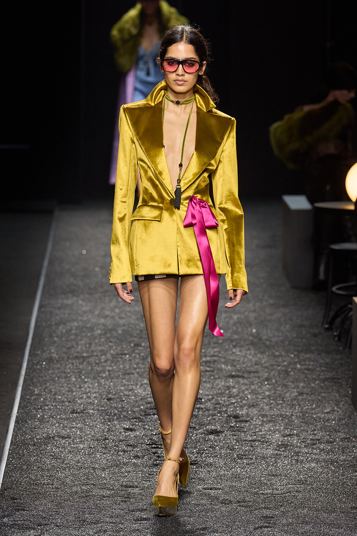 Model walks the runway in a metallic gold satin blazer mini dress with a bright pink ribbon belt and pink sunglasses, heels included.