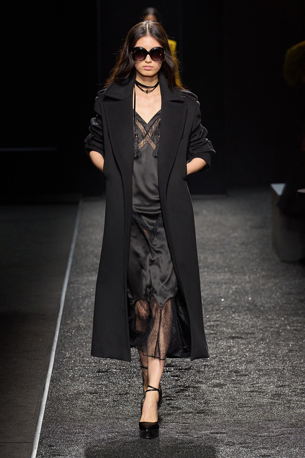 Model on a runway wearing a long black coat over a sheer black lace dress and black heels, with oversized sunglasses.