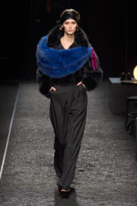 Model walking a runway in an oversized black fur coat with a bright blue fluffy scarf draped over the shoulders.