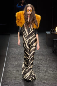 Model walking runway in a beige-black zebra-print jumpsuit with orange faux-fur shoulder accents and yellow-tinted sunglasses, black gloves.