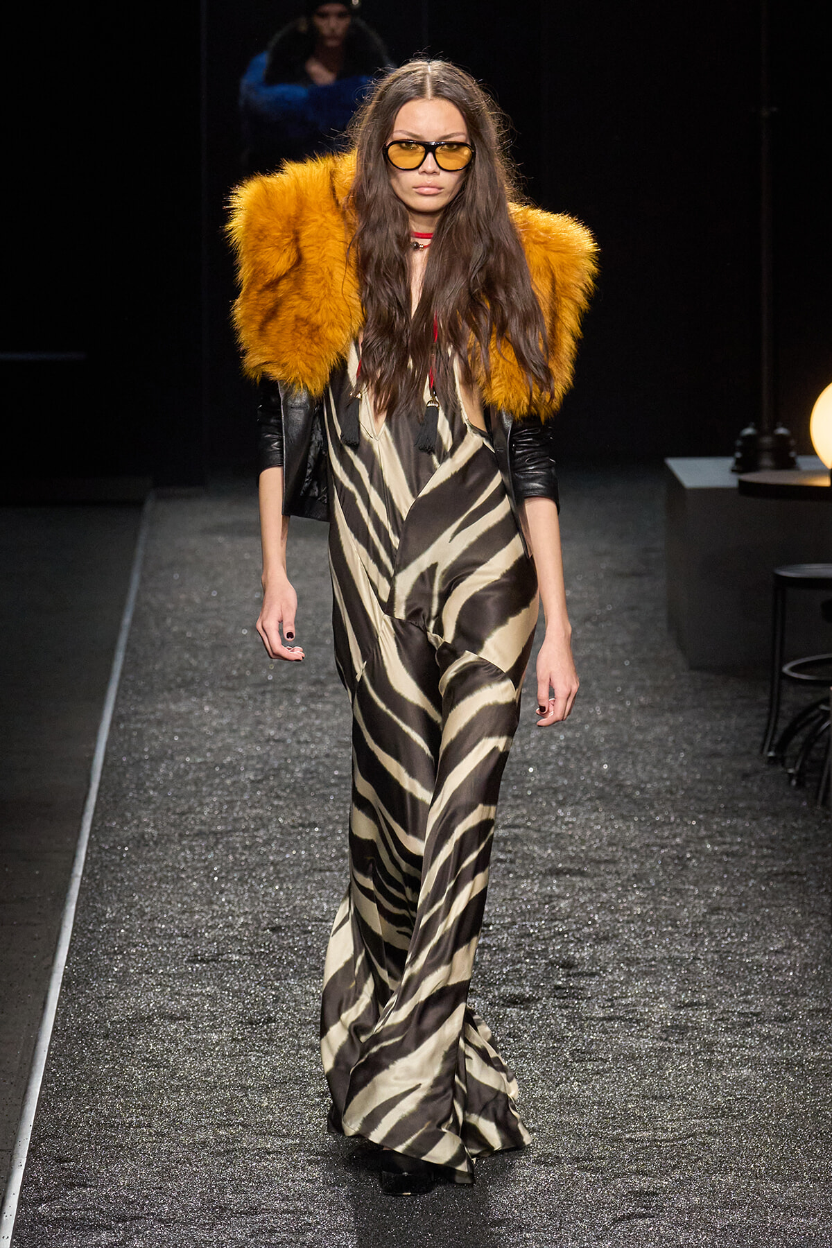 Model walking runway in a beige-black zebra-print jumpsuit with orange faux-fur shoulder accents and yellow-tinted sunglasses, black gloves.