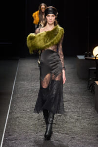Model on a glittery runway wearing a black lace gown with sheer panels and a lime-green fur stole draped across one shoulder.