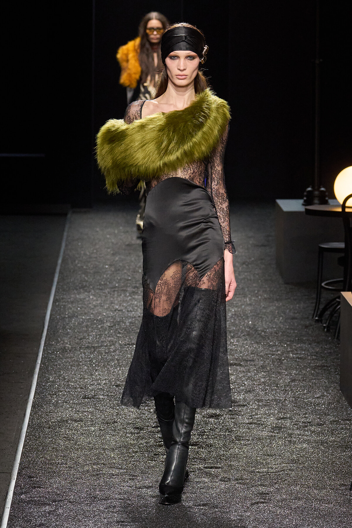 Model on a glittery runway wearing a black lace gown with sheer panels and a lime-green fur stole draped across one shoulder.
