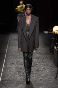 Model on a dim runway wearing a dark oversized blazer, sheer black top, blue strap necklaces, and black thigh-high boots.