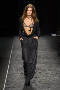 Female model walks a runway in a black sheer lace bralette, draped top, and pinstripe wide-leg pants, with layered necklaces.