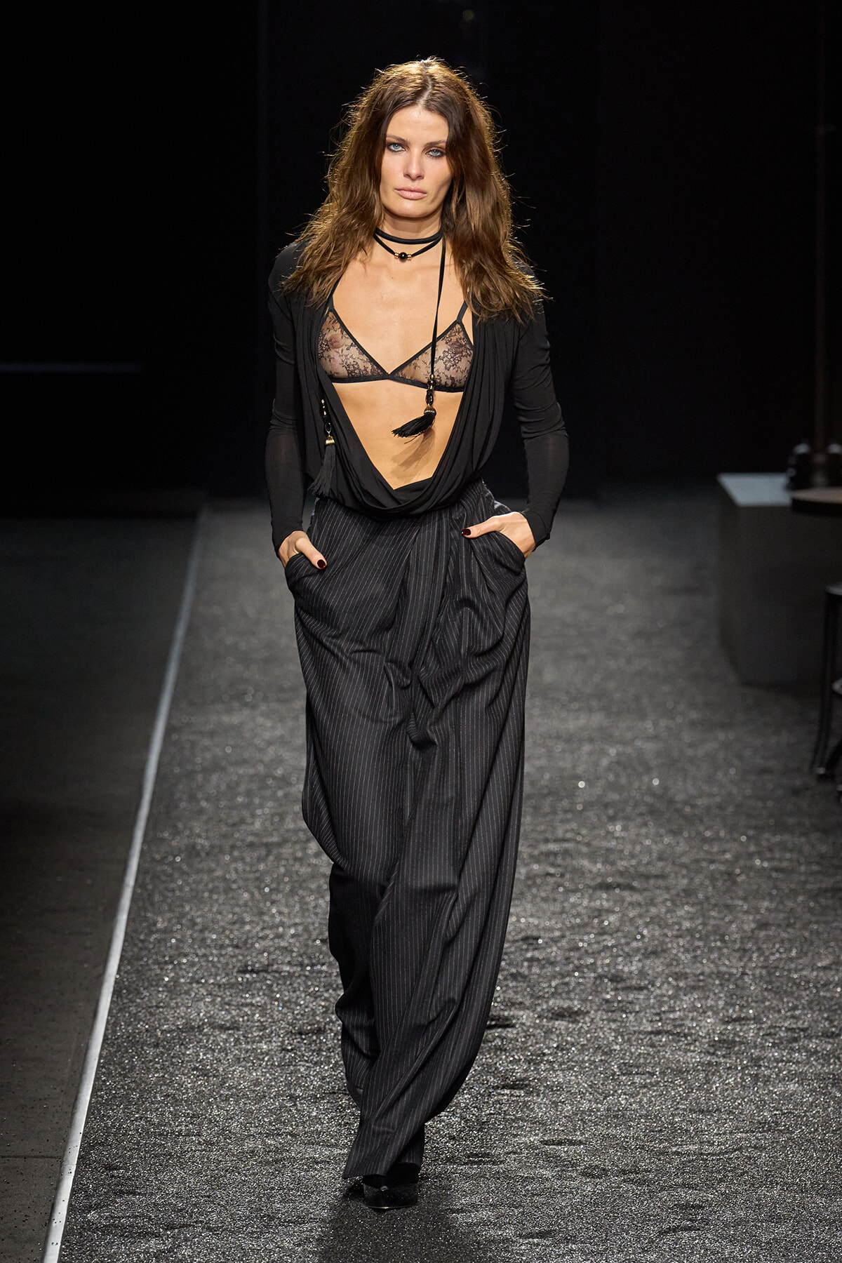 Female model walks a runway in a black sheer lace bralette, draped top, and pinstripe wide-leg pants, with layered necklaces.