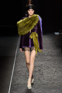Model walking a fashion runway in a purple velvet blazer, lime-green fur stole, and a wide yellow ribbon belt over lace-trim shorts, beige heels.