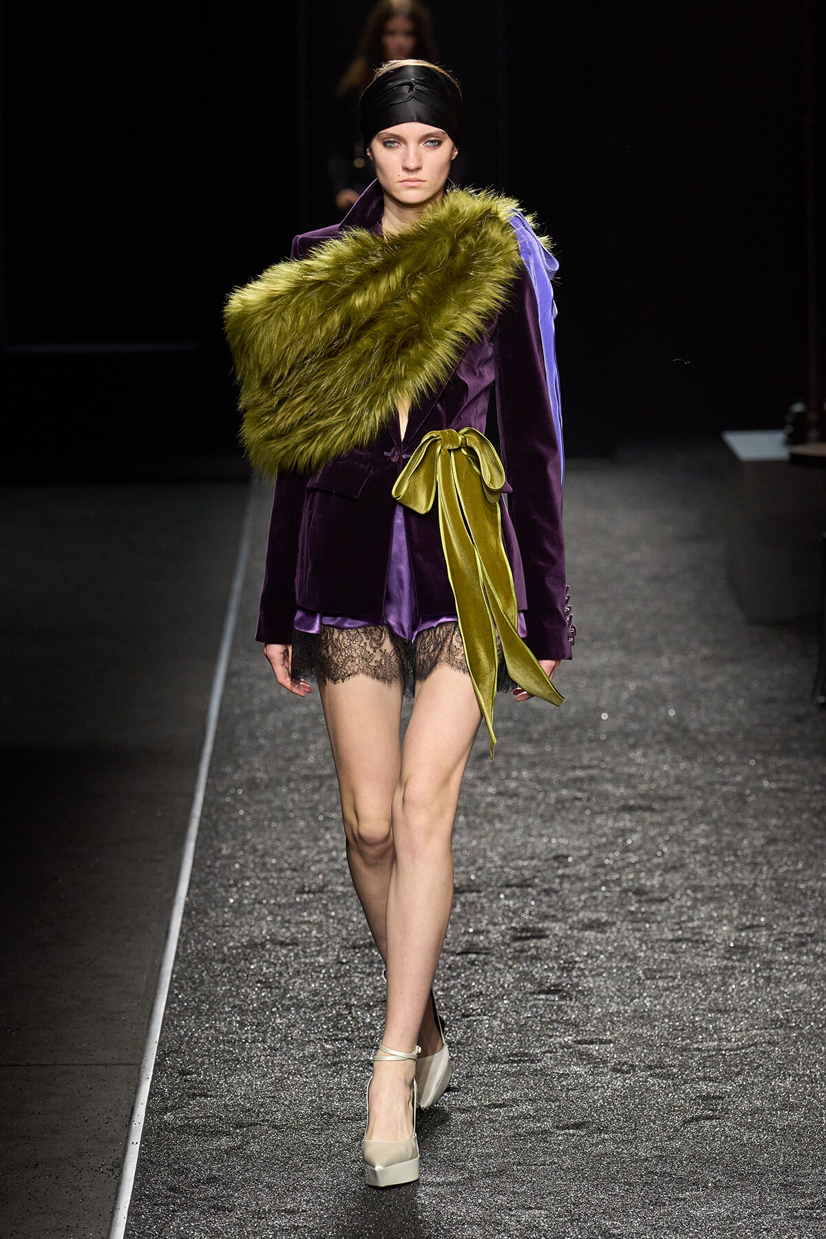 Model walking a fashion runway in a purple velvet blazer, lime-green fur stole, and a wide yellow ribbon belt over lace-trim shorts, beige heels.