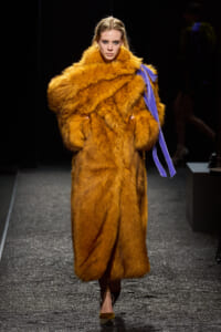 Model walks the runway wearing a voluminous orange fur coat with a purple sash draped over the shoulder.