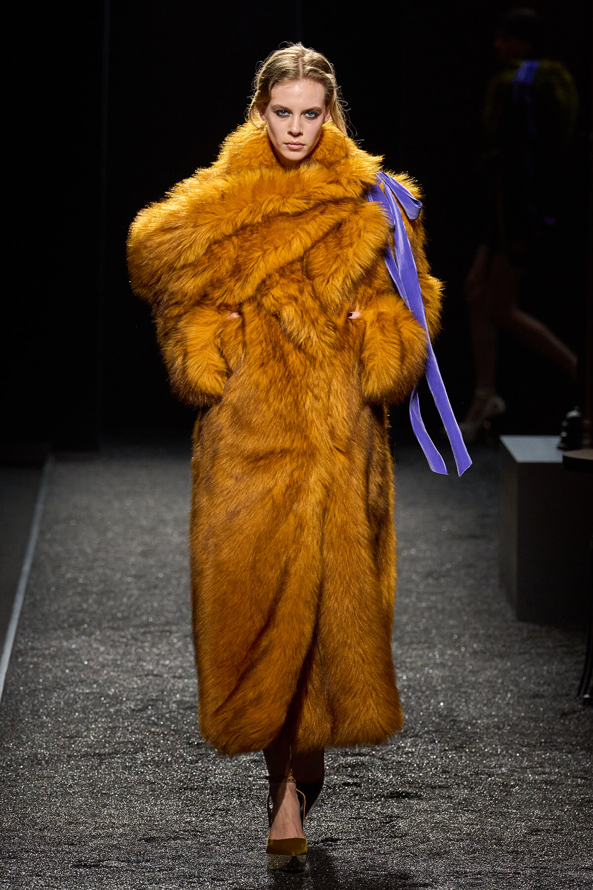 Model walks the runway wearing a voluminous orange fur coat with a purple sash draped over the shoulder.