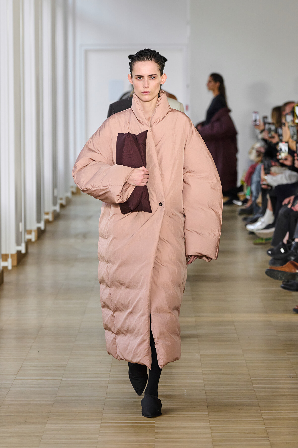 Model walking a runway in a pale pink oversized puffer coat, holding a dark burgundy clutch; audience with phones in the background.