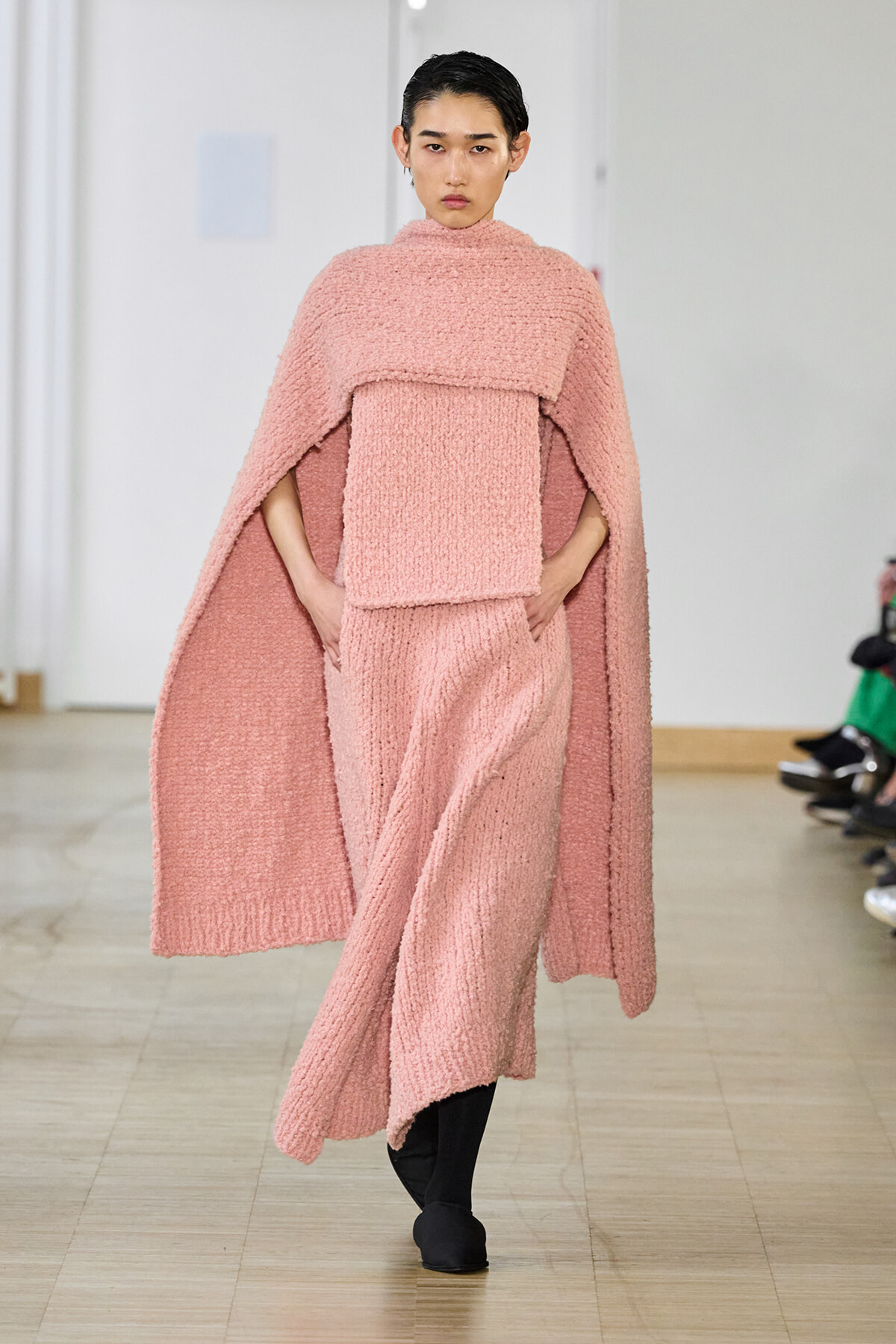 Model walks runway in a soft pink textured cape over a matching knit dress, hands in pockets.