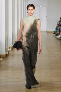 Model walks a fashion runway in a sleeveless, gradient jumpsuit with beige to dark brown splatter pattern, holding a black clutch.