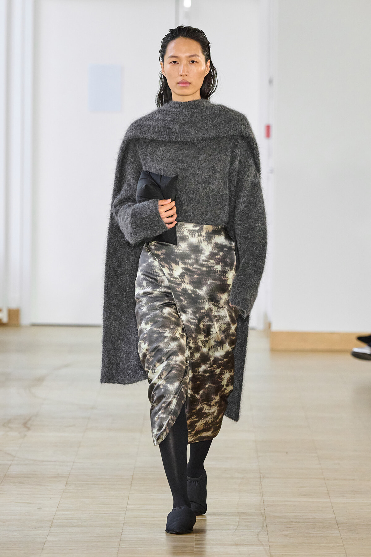 Model walking runway in a dark gray oversized fuzzy sweater and metallic camouflage wrap skirt, holding a black clutch.