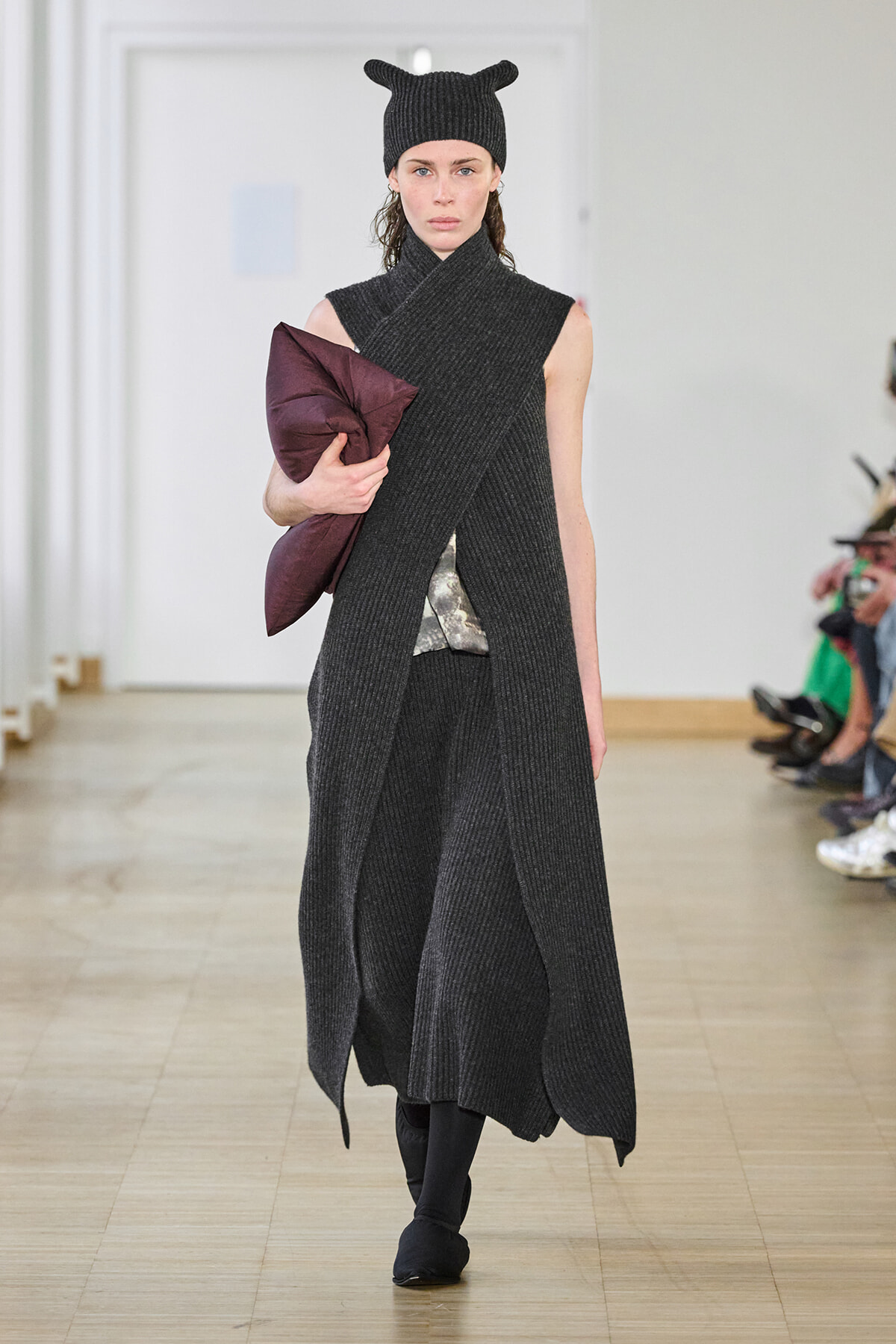 Model walks runway in a long charcoal knit wrap coat and matching hat, holding a burgundy clutch.