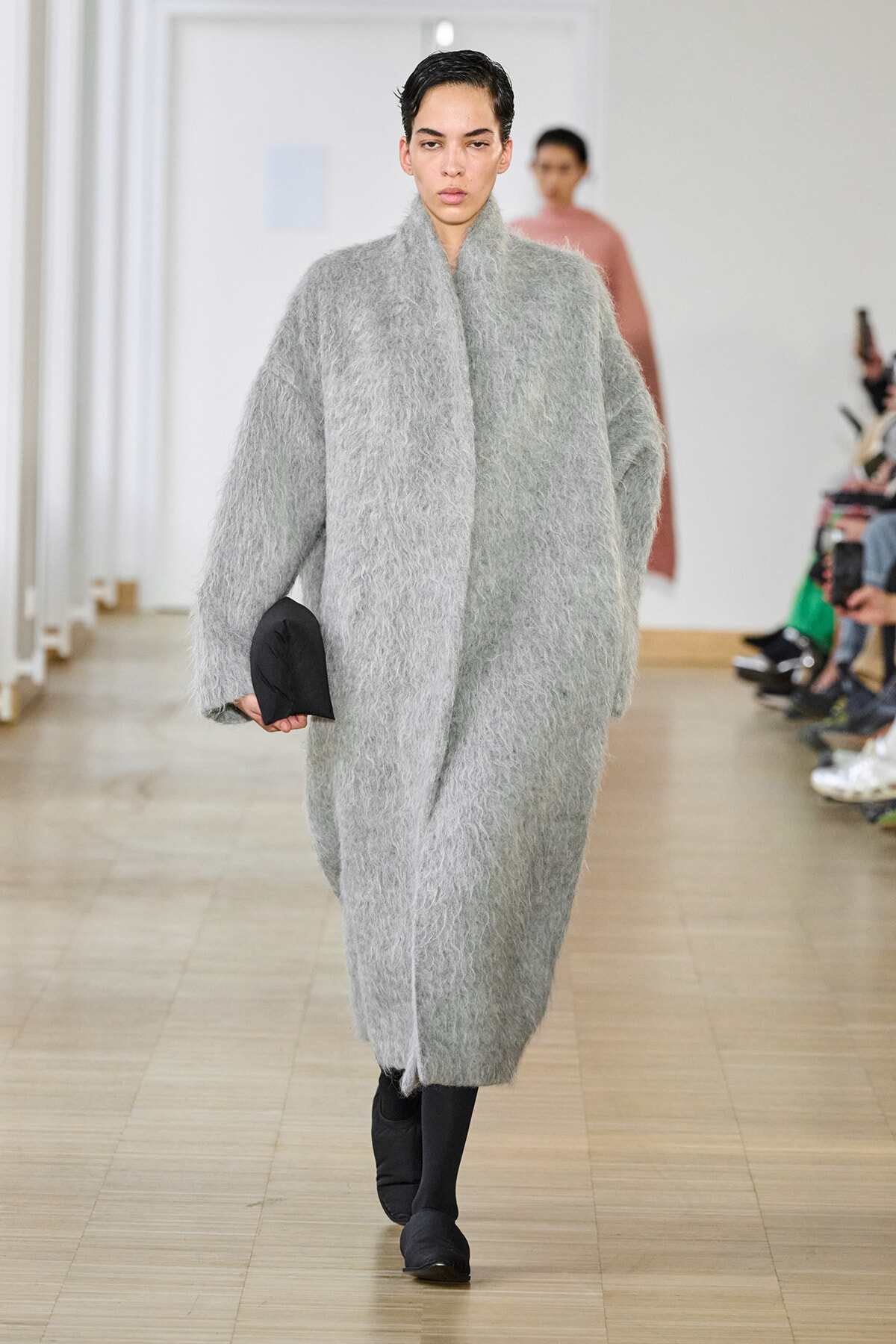Model walks the runway in a bulky, light-gray textured coat with black shoes and a small black clutch in her hand.