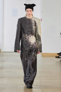 Model on a runway wearing a grayscale splatter-print tailored top and wide-leg pants with a black knit beanie.