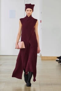 Model wearing a burgundy knit wrap dress and matching hat, carrying a pale pink clutch on the runway.