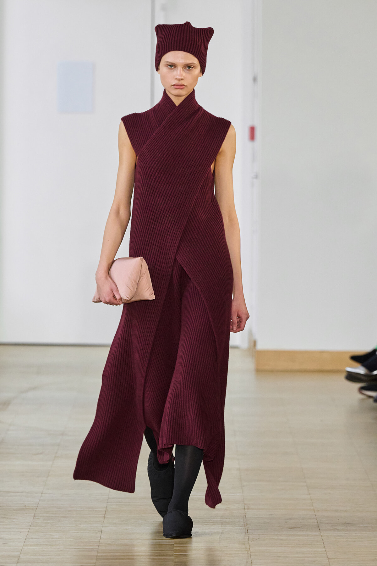 Model wearing a burgundy knit wrap dress and matching hat, carrying a pale pink clutch on the runway.