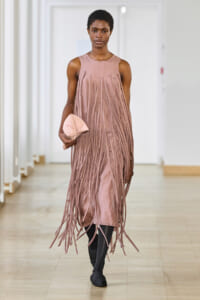 Model walking a runway in a sleeveless dusty-pink fringe dress, holding a matching pink clutch.