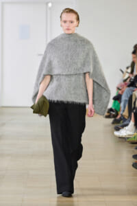 Model walking the runway in a bulky light-grey fuzzy poncho with a high neck over wide-leg black pants, holding an olive clutchShifted to her side.