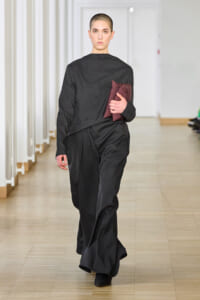 Female model with a shaved head wearing a black loose-fitting outfit, walking down a runway while holding a burgundy clutch.