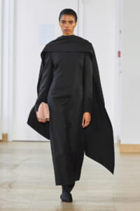 Model wearing a black cape-style coat over a black jumpsuit, holding a pale pink clutch.