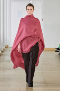 Model walks the runway wearing a loose pink-red cape draped over black trousers in a fashion show venue.