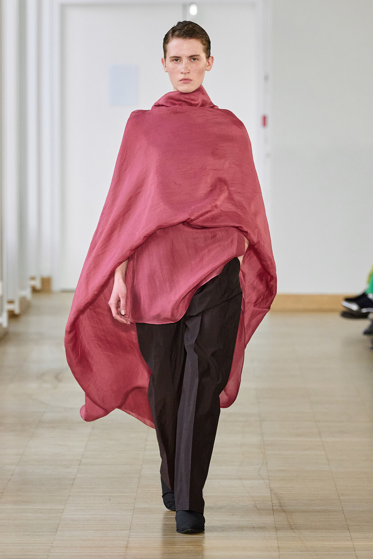 Model walks the runway wearing a loose pink-red cape draped over black trousers in a fashion show venue.