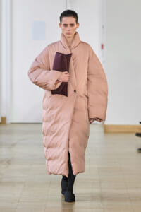 Model on a runway wearing a long oversized pale pink puffer coat, holding a dark burgundy clutch under her arm.