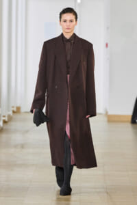 Model walks a runway in a long brown overcoat over a dark outfit, holding a black clutch.