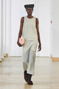 Model walking a runway in a pale beige sleeveless tunic and wide-leg pants, black knit turban, holding a pink clutch, wearing dark socks and brown fuzzy slippers.