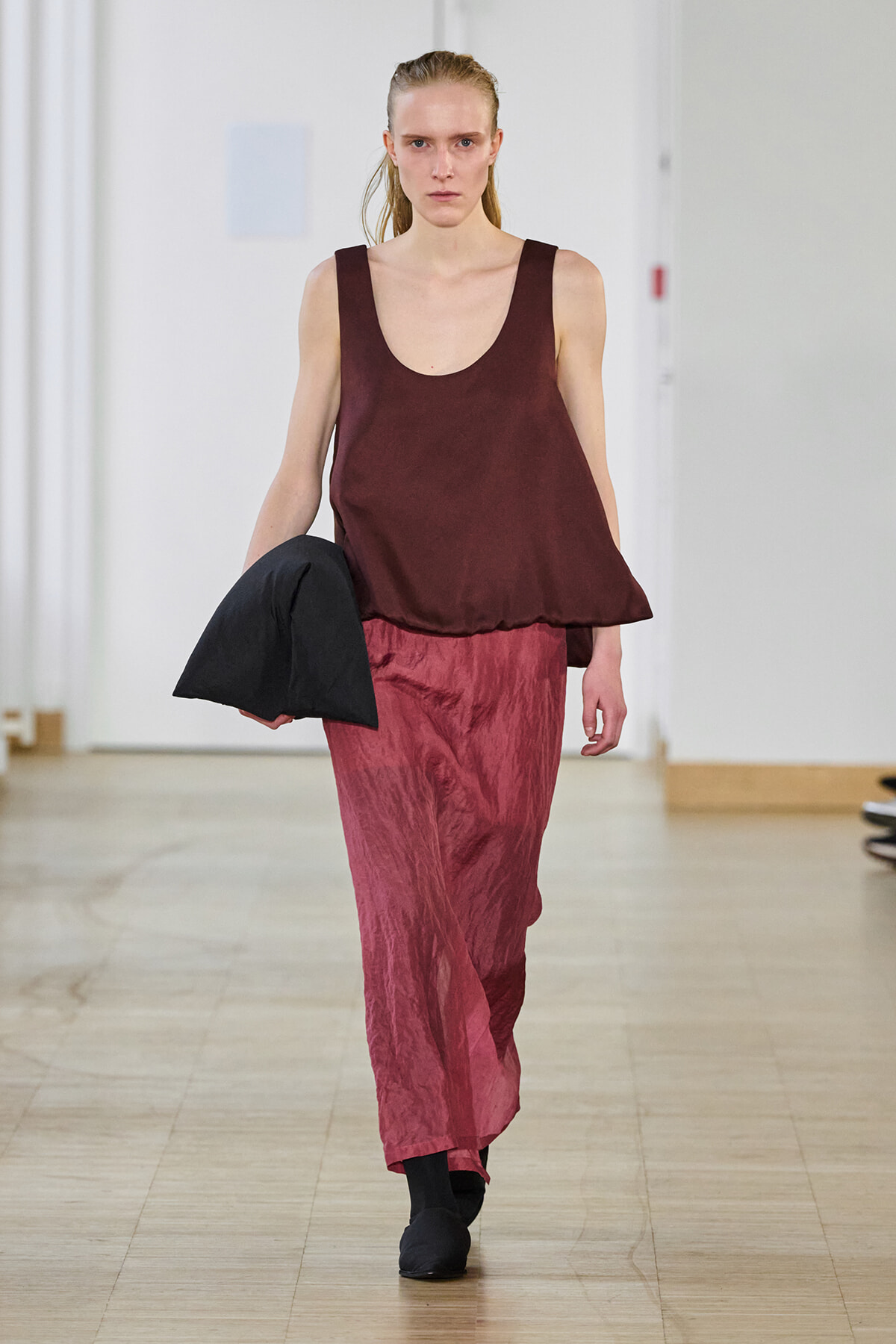 Runway model walks in a burgundy sleeveless top with crinkled pink pants, holding a black foldover bag sidewarded.