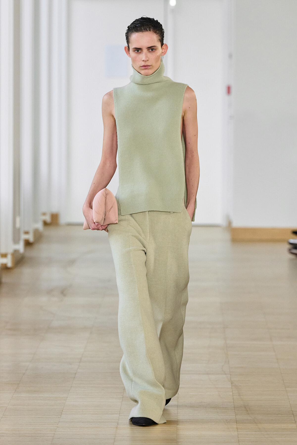 Runway model in a mint sleeveless turtleneck top and matching wide-leg pants, holding a pale pink clutch in her left hand.
