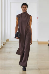 Model walking down a bright hallway in a sleeveless brown high-neck dress, holding a small dark clutch.