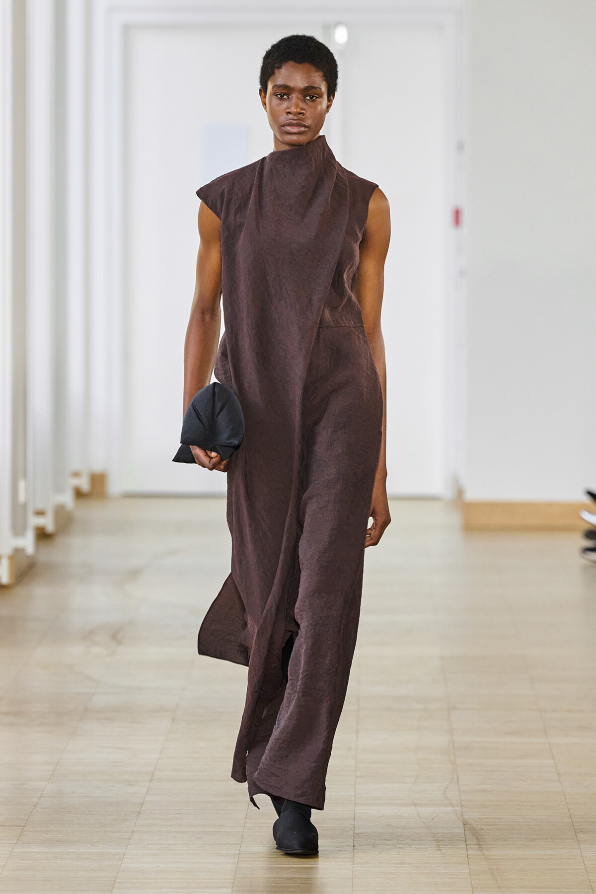 Model walking down a bright hallway in a sleeveless brown high-neck dress, holding a small dark clutch.
