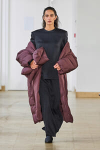 Model walking down a minimalist runway in a dark, structured dress accented by a oversized maroon puffer coat draped over shoulders.