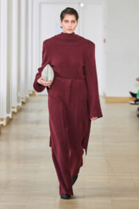 Model walking a runway in a burgundy oversized wool coat, holding a light gray clutch at waist level.
