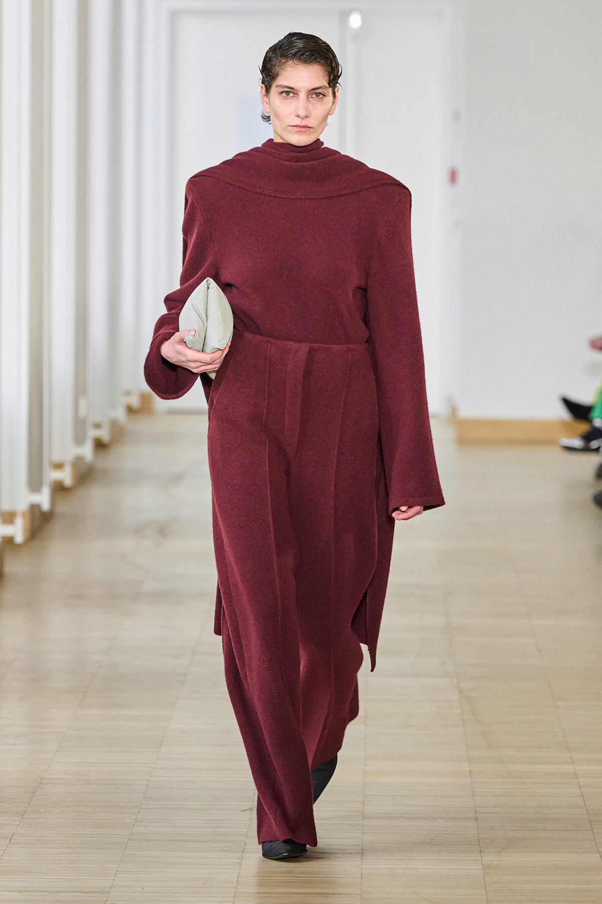 Model walking a runway in a burgundy oversized wool coat, holding a light gray clutch at waist level.