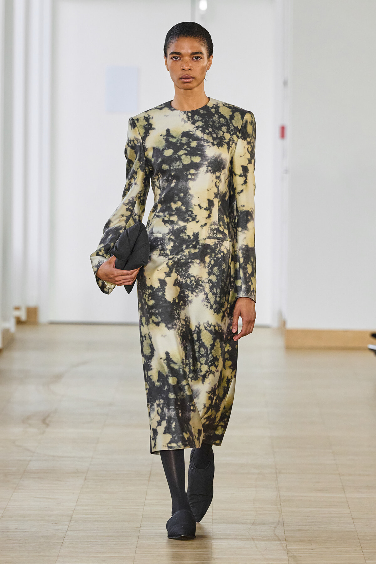 Model walking a runway in a long-sleeve beige and black tie-dye midi dress, carrying a black clutch and wearing dark ankle boots.