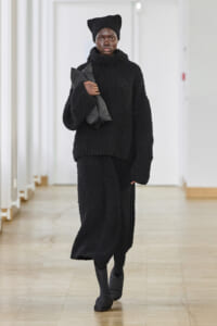 Model wearing a black textured knit ensemble with a beanie, walking down a hallway while holding a folded gray garment under her arm.