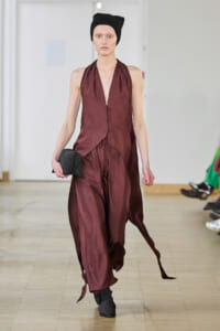 Model walks a runway wearing a burgundy satin halter jumpsuit with flowing draped panels, black beanie, and black shoes, holding a dark cap.