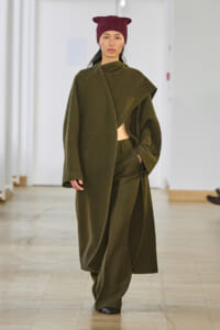 Model walking a runway in an oversized olive-green draped coat with matching wide-leg pants, wearing a burgundy knit beanie.