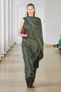 Model walking a runway in a loose, olive-green draped gown, holding a maroon rounded clutch.