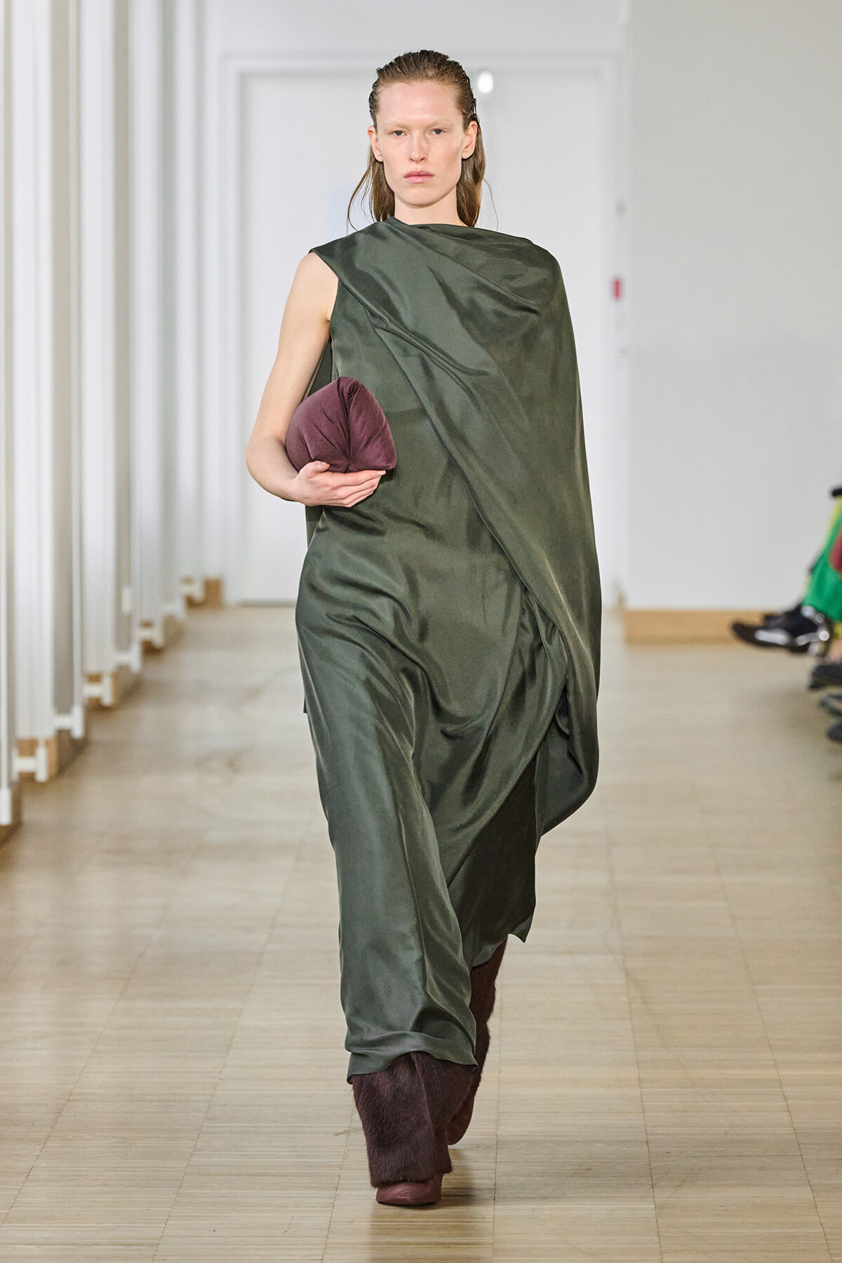 Model walking a runway in a loose, olive-green draped gown, holding a maroon rounded clutch.