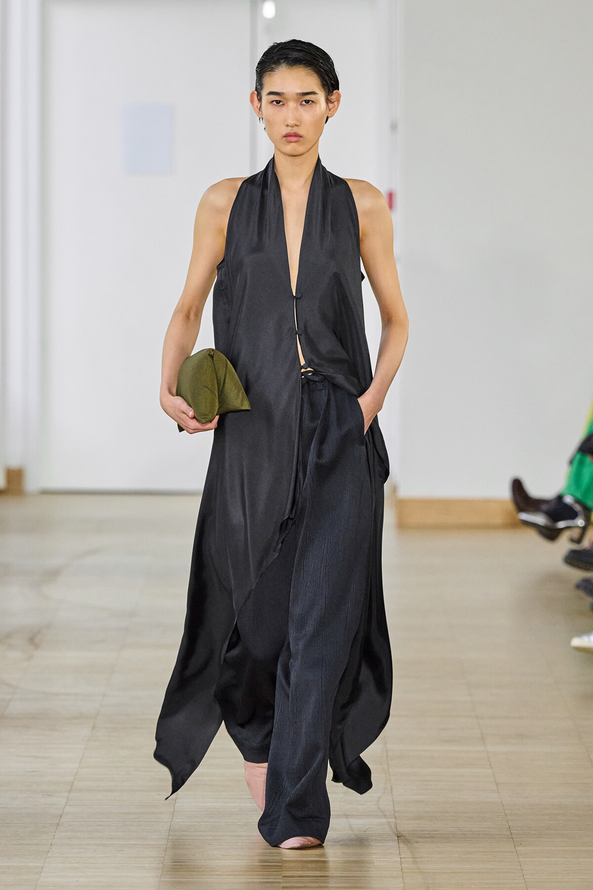 Runway model in a sleeveless black halter dress with a plunging neckline, holding an olive-green clutch in one hand.