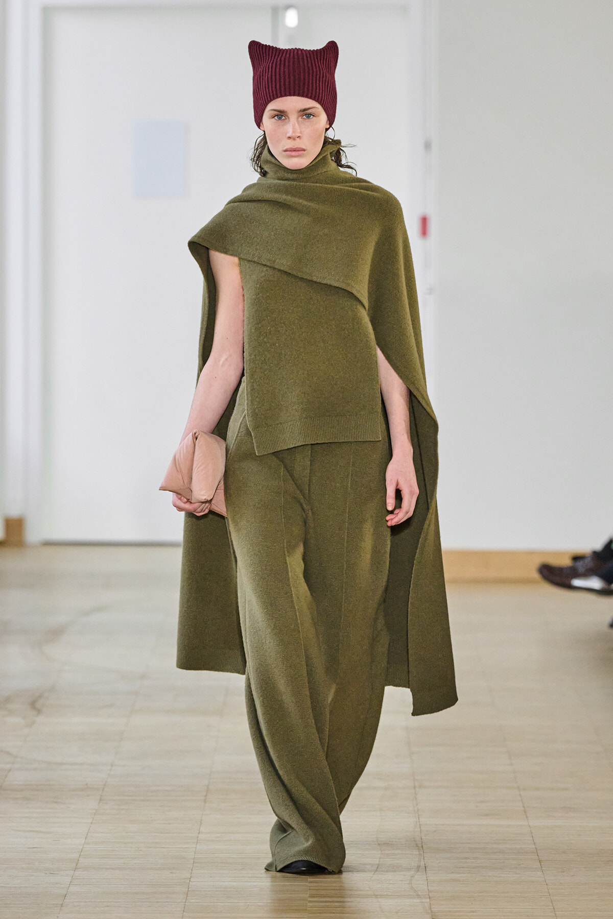 Model on a runway in an olive-green cape and matching wide-leg pants, paired with a burgundy knit hat and beige clutch.