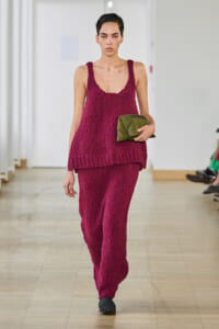 Model walks a runway in a textured magenta knit sleeveless top and matching pants, carrying an olive-green clutch.