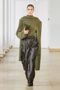 Model with a shaved head walks a runway wearing an olive green oversized sweater and black shiny leather pants, clutch in hand in a neutral hallway setting.
